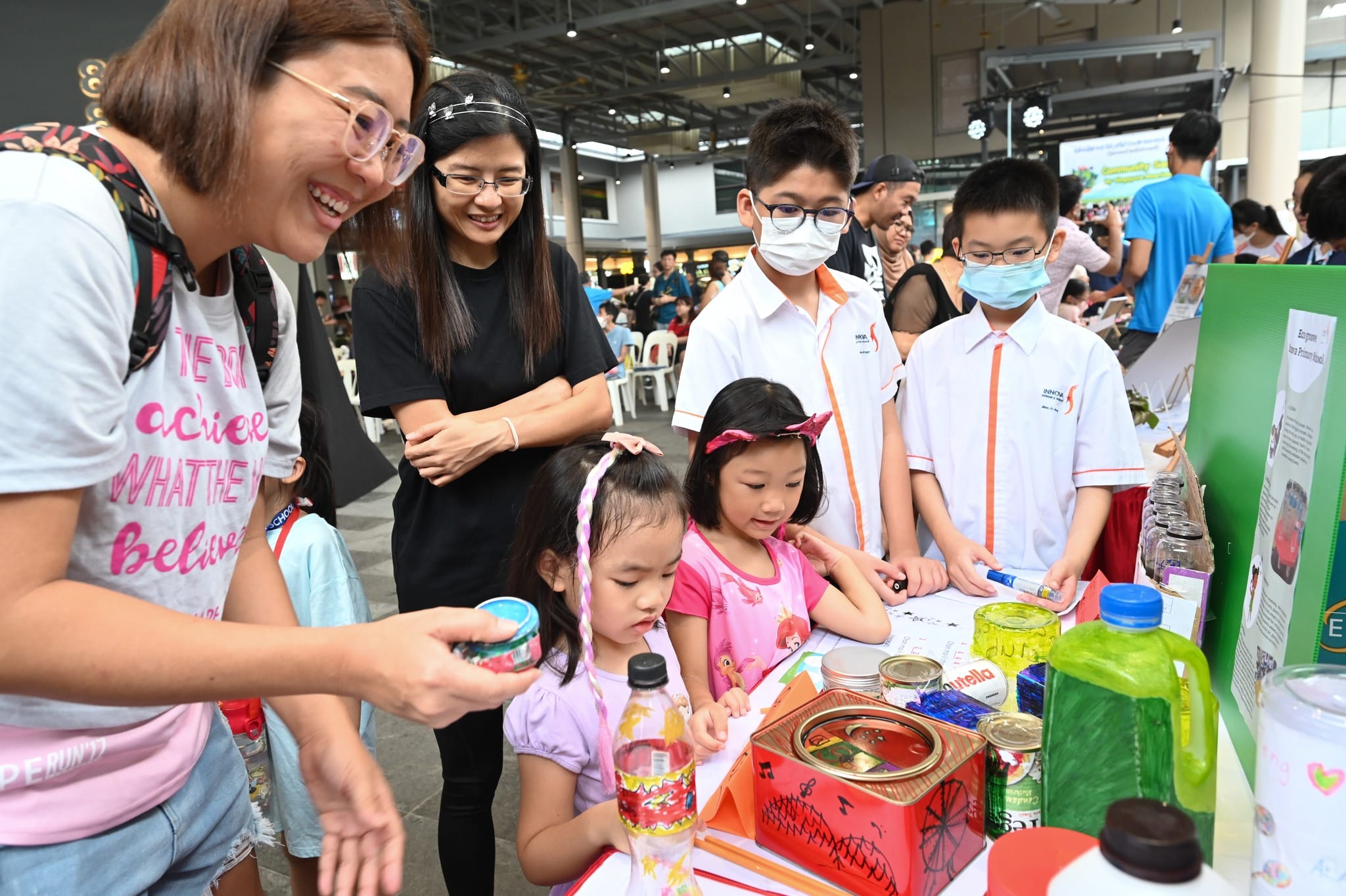 Children and adults gather around a display table with bottles and cans at an indoor community fair event.